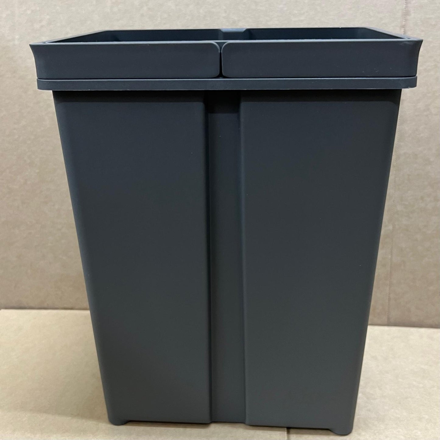 Dark gray recycling bin with two compartments on a beige floor.